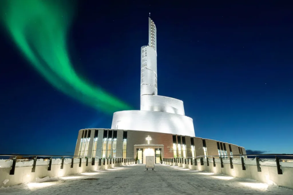 Northern Lights Cathedral in Alta, Norway with the Cross of Christ light in bright white with northern lights dancing above the cathedral in the sky representing captivating norther lights cruise destinations offered by MSC, Holland America, Princess Cruises and Norwegian Cruise Lines.
