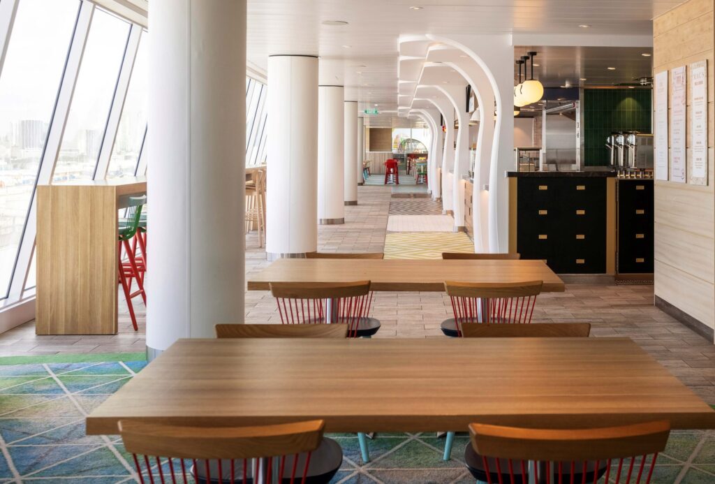 Hall view of the Icon ship Aqua Dome Market Side eatery table area with window views and buffet style chiefs' area representing endless all you can eat dining experiences while you cruise into your dream vacation.